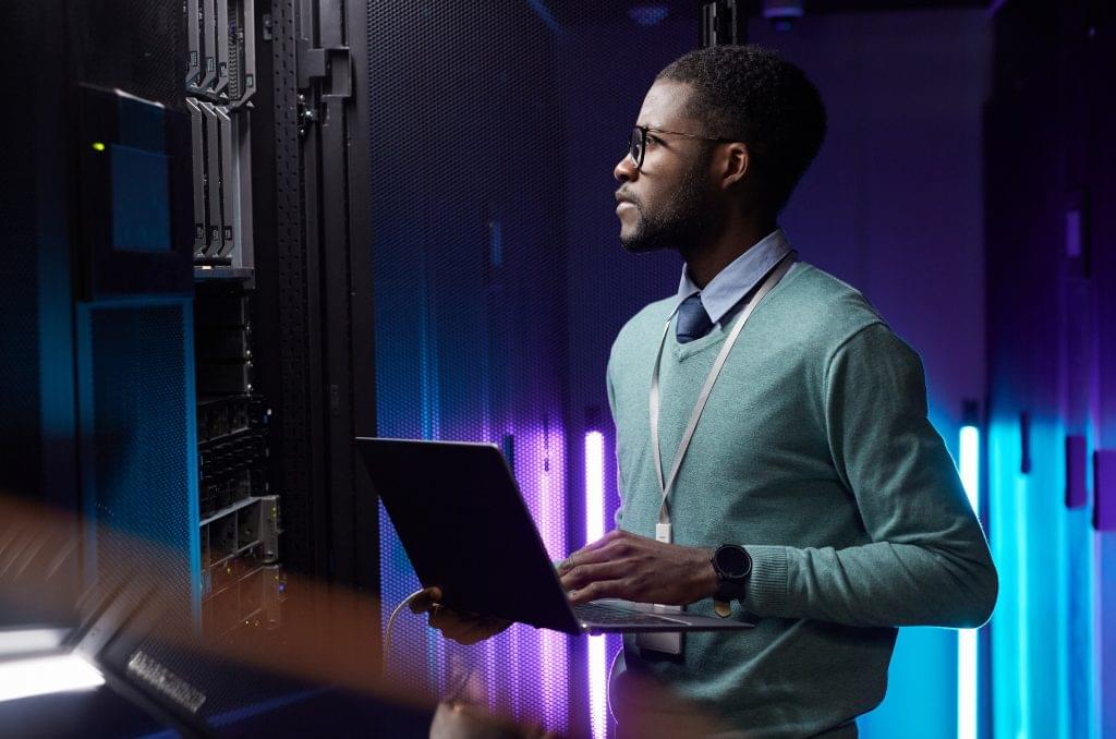 Data engineer holding laptop while working in server room lit by blue light