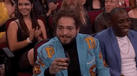 Post Malone, glass of wine in hand, grins and raises his eyebrows at the camera