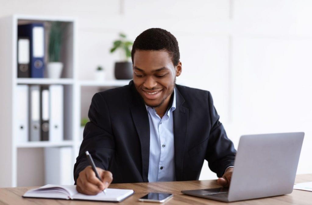 Millennial man taking notes at a laptop with a cellphone on the desk.