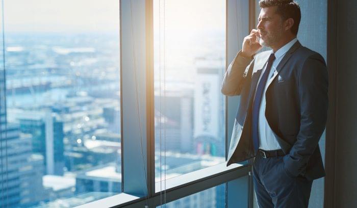 Business executive with designer stubble, talking confidently on his mobile phone while looking out of large windows in a top floor office at the city below